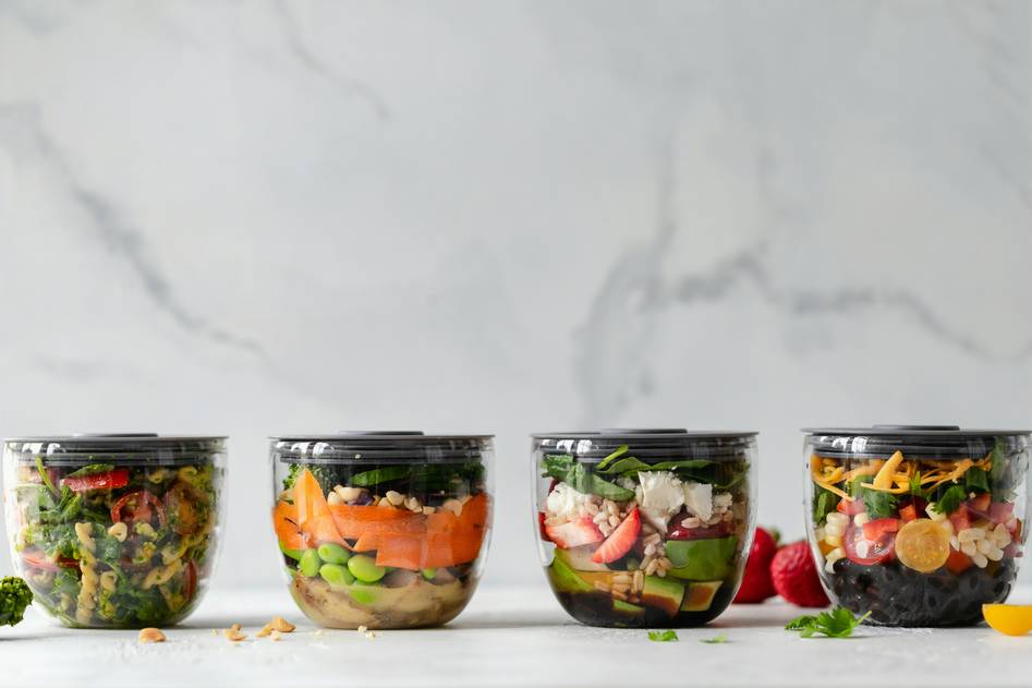 Two hands holding open a set of colorful, stackable reusable snack containers on a rustic kitchen table with herbs in the background